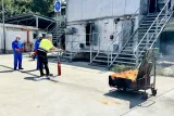 Гасене на горяща тава с пожарогасител; двама курсисти с каски наблюдават пред огневата сграда.